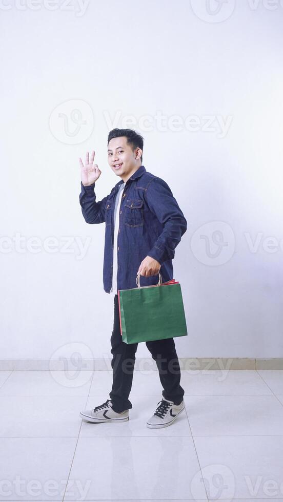 Portrait full body of Asian man holding shopping bags and showing OK sign gesture with hand isolated on white background photo