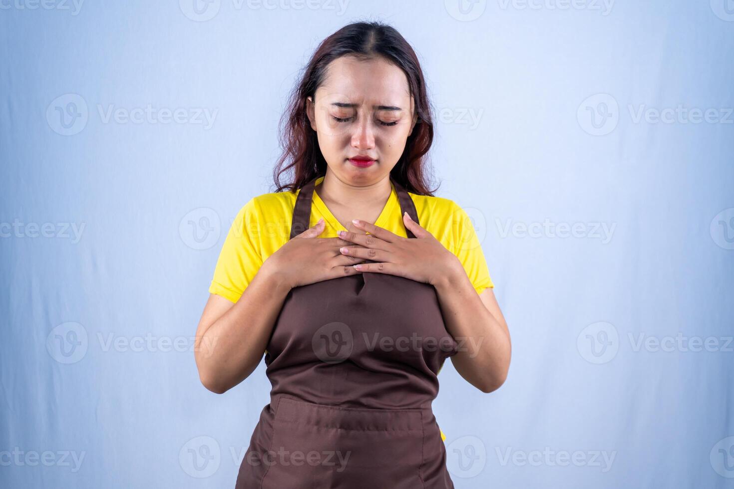 Sad Asian woman in yellow shirt and apron, eyes downcast, hands on chest, troubled. photo
