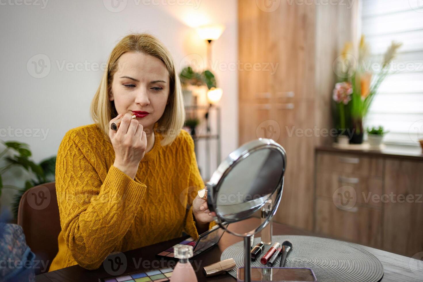 Young woman applying lipstick while seated at a stylish vanity in a cozy modern room with warm lighting photo