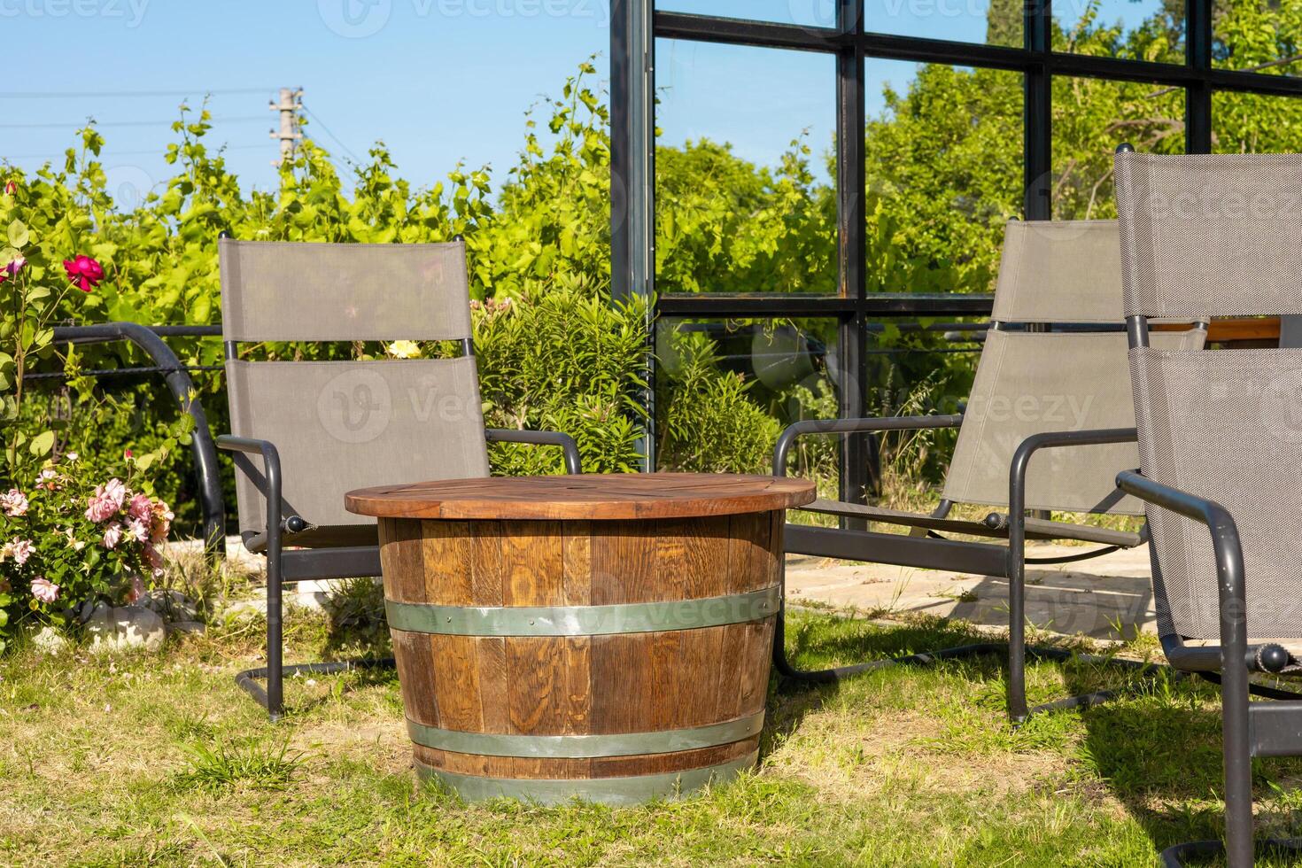 A rustic wooden barrel table sits on green grass, surrounded by two modern gray lounge chairs. Lush greenery and flowers create serene outdoor setting in Turkiye. photo