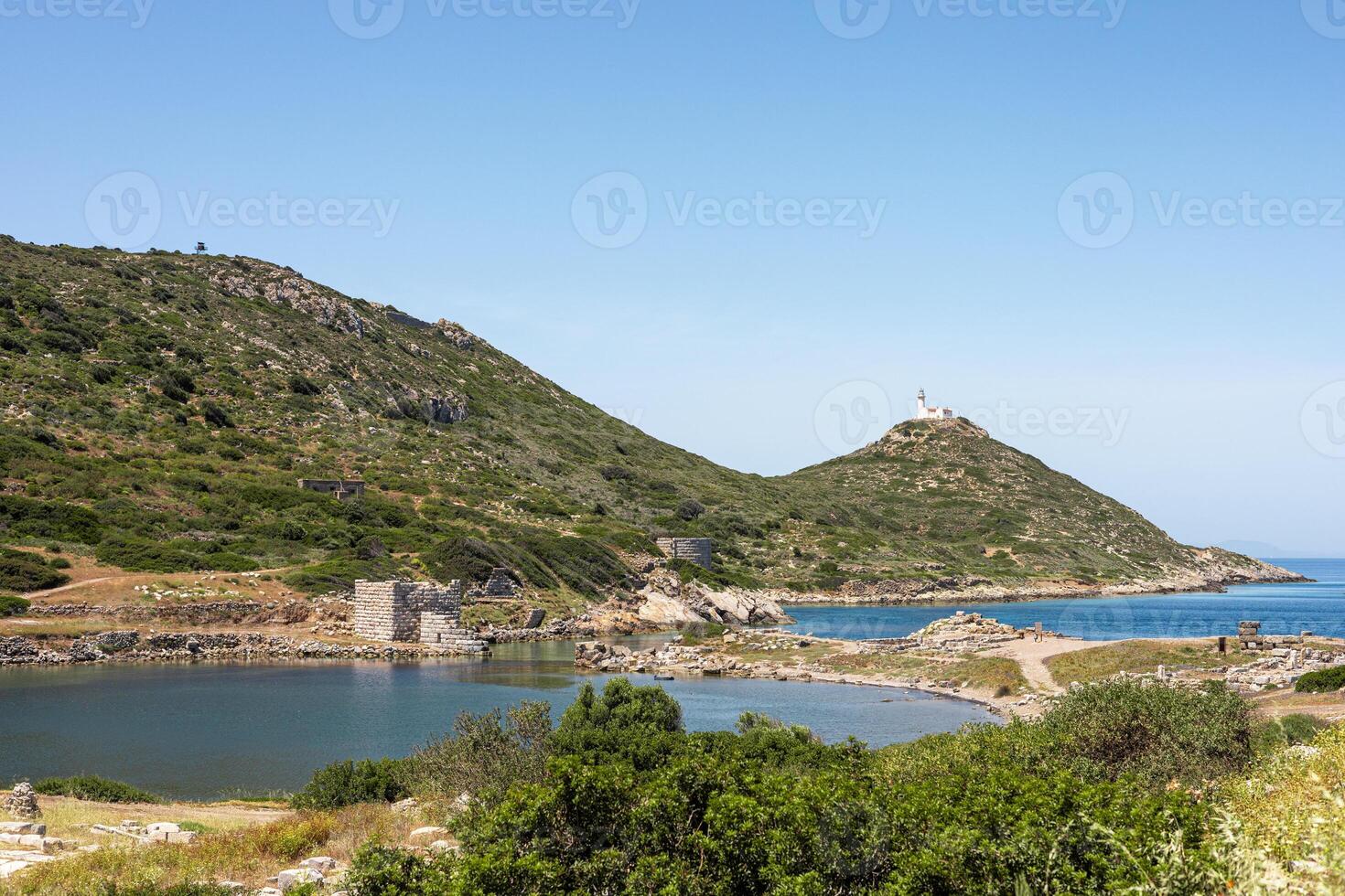 A scenic view of a coastal landscape in Turkiye. Green hills rise above a calm blue sea, with a small harbor and distant lighthouse visible under a clear sky. photo