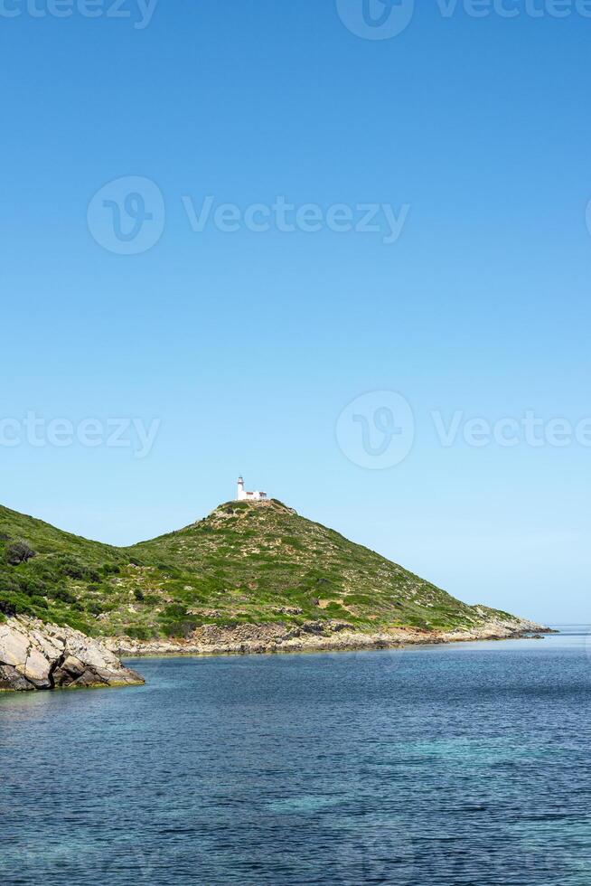 A serene coastal view of a green hill in Turkiye, with a lighthouse at the top, under a clear blue sky and calm water photo