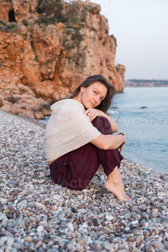 A young Caucasian woman with brown hair sits on a pebbly beach by the sea. She wears a light shawl and dark pants, with rocky cliffs in background. photo