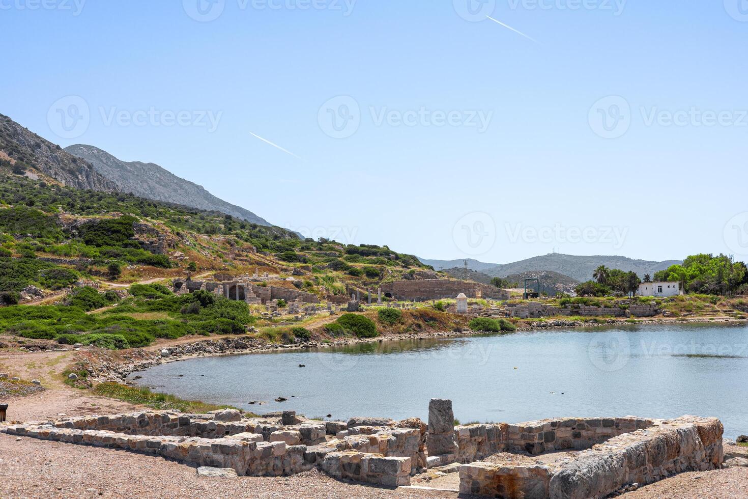 A scenic view of ancient ruins by a calm lake in Turkiye. Lush green hills surround the area, with mountains in the background under a clear blue sky. photo