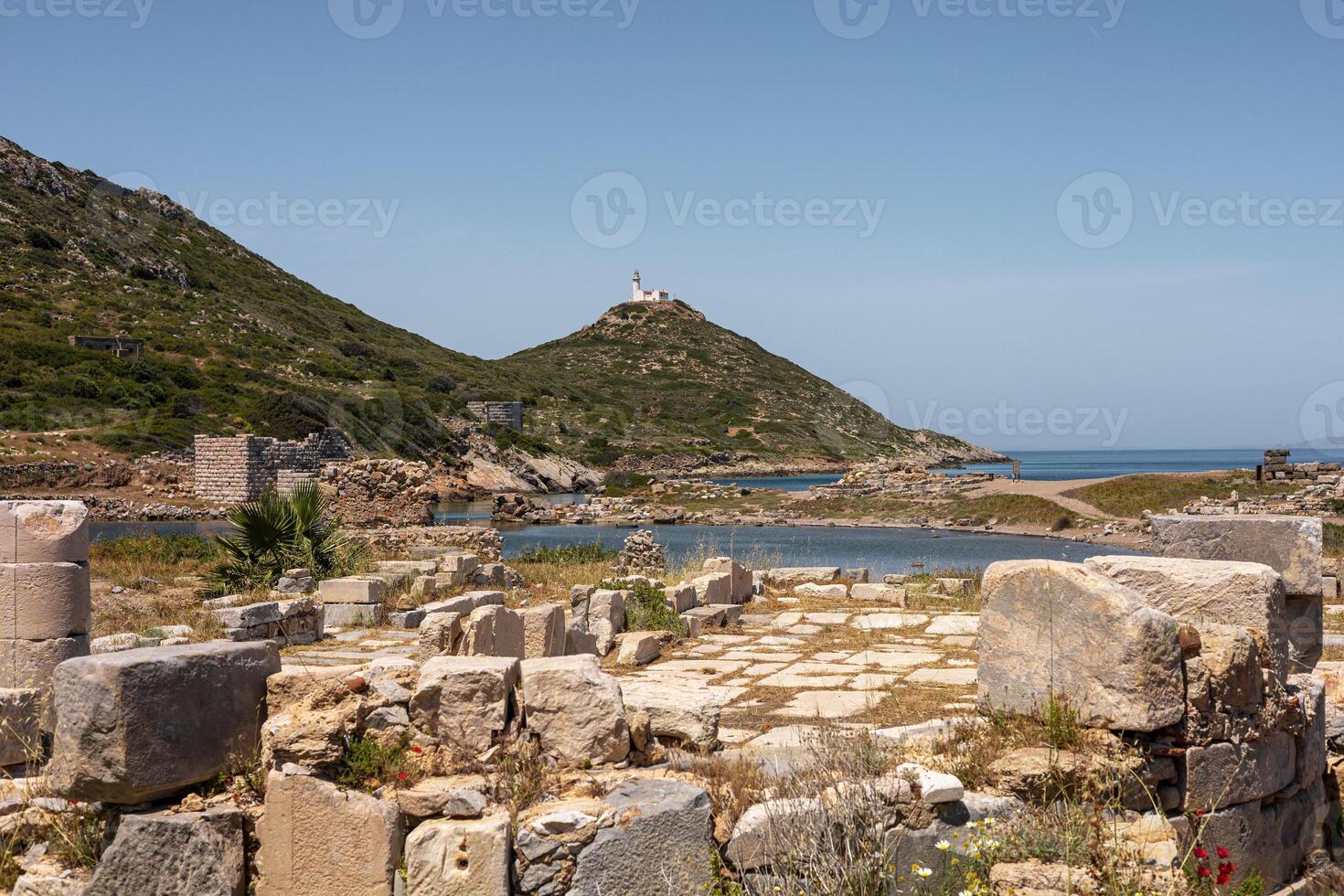Ancient ruins near the coast in Turkiye. Stone structures and columns are visible. Green hills rise in the background, leading to a lighthouse on a hilltop. photo