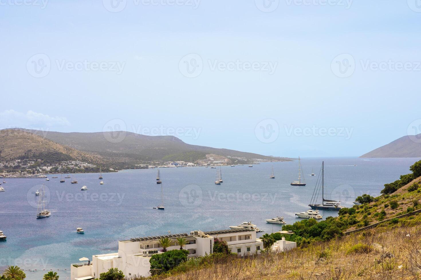 A scenic view of a calm bay with several boats anchored. Rolling hills and a clear sky create a tranquil atmosphere. White buildings line the shore. photo