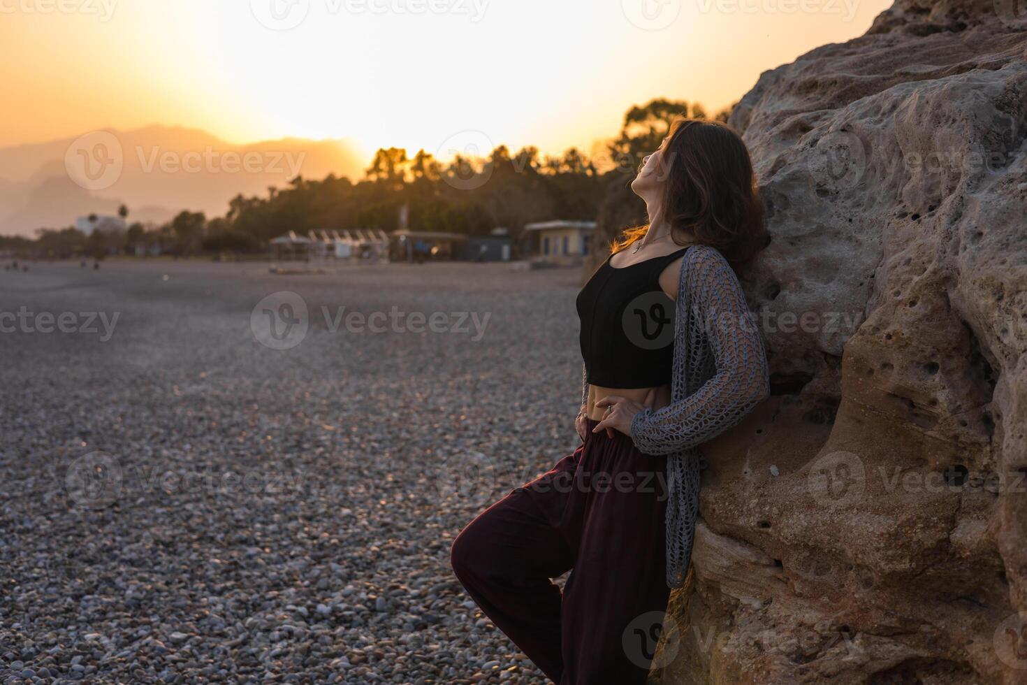 A young Caucasian woman with long brown hair stands by rocky beach at sunset. She wears a black top and maroon pants, enjoying the serene atmosphere. photo