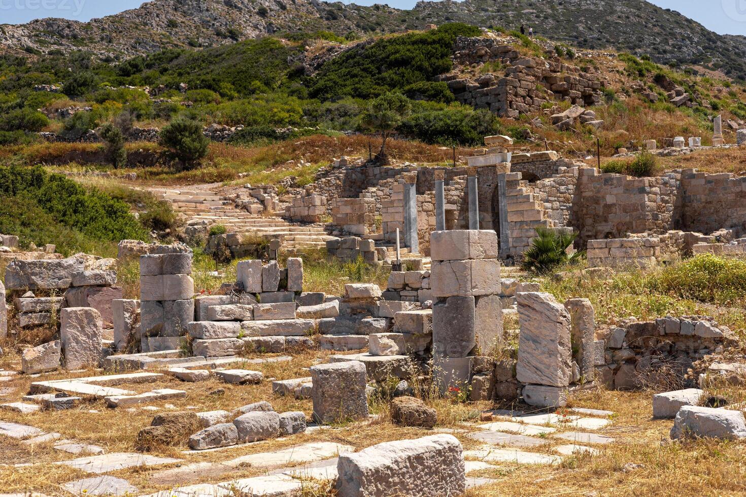 Ancient ruins in Turkiye with stone columns and pathways. Lush greenery surrounds the archaeological site, set against a mountainous backdrop. photo