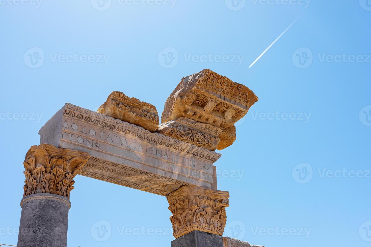 Ancient stone ruins with ornate columns under a clear blue sky. A jet trail is visible in the background, highlighting the historical significance of the site. photo