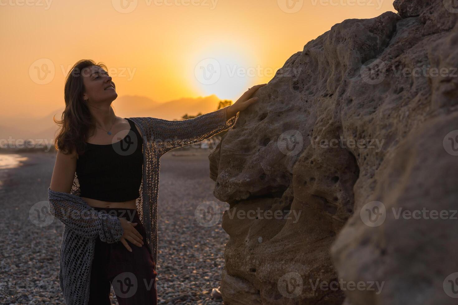 A young Caucasian woman with long brown hair stands by a rocky beach at sunset. She wears a black top and maroon pants, enjoying serene atmosphere photo