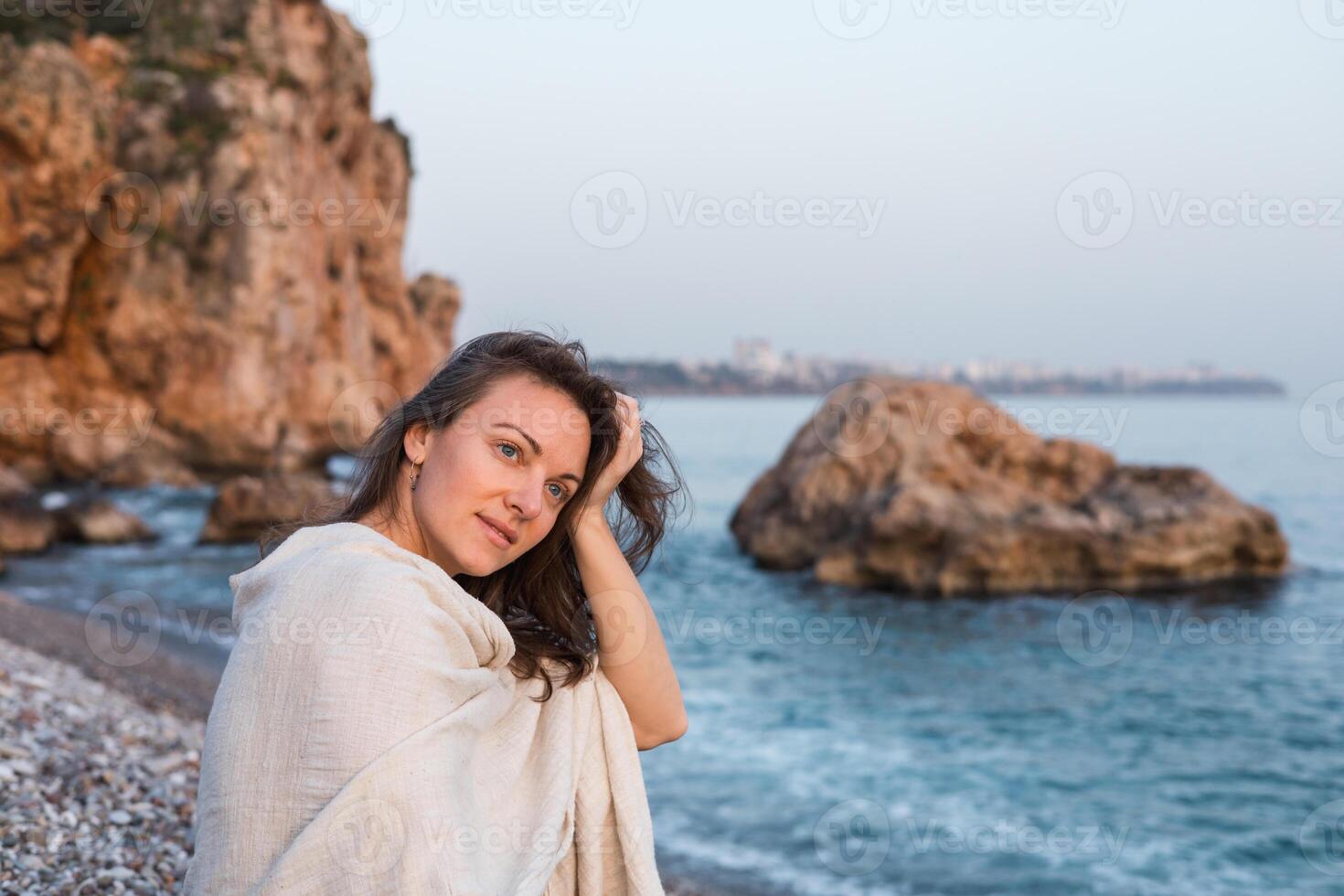 A young Caucasian woman with long brown hair poses on a beach near rocky cliffs. The ocean is calm, and the sky is clear during sunset. photo