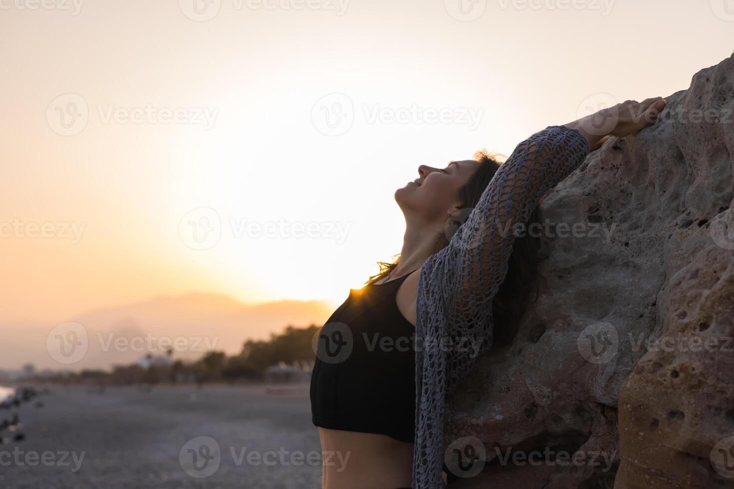 A young Caucasian woman with brown hair climbs a rocky formation at sunset. The sun sets behind her, casting warm glow over beach scene. photo