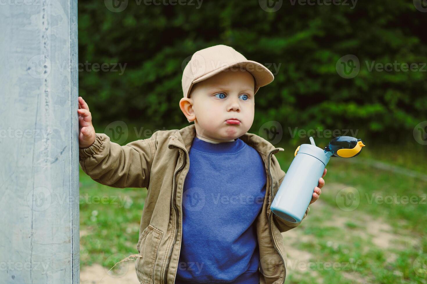 A delightful and cute toddler is enjoying the outdoors while playfully holding a water bottle and wearing a cap, surrounded by a vibrant and cheerful natural setting, filled with greenery and joy photo