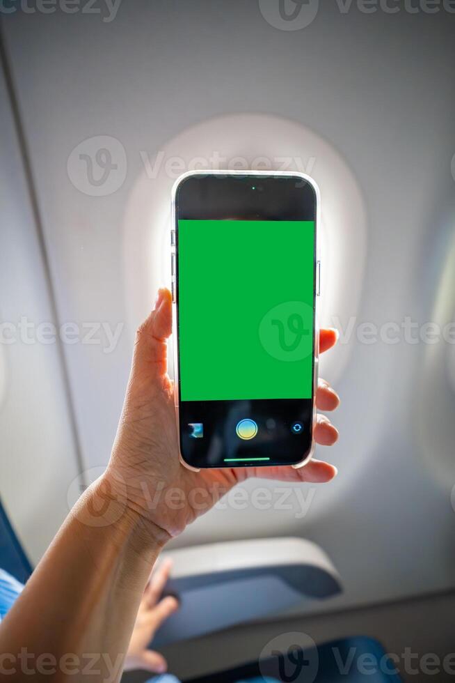 Close-up of a hand holding a smartphone with green screen camera active, recording view from airplane window. Travel content creation, mobile photography, and mockup preparation in flight. photo