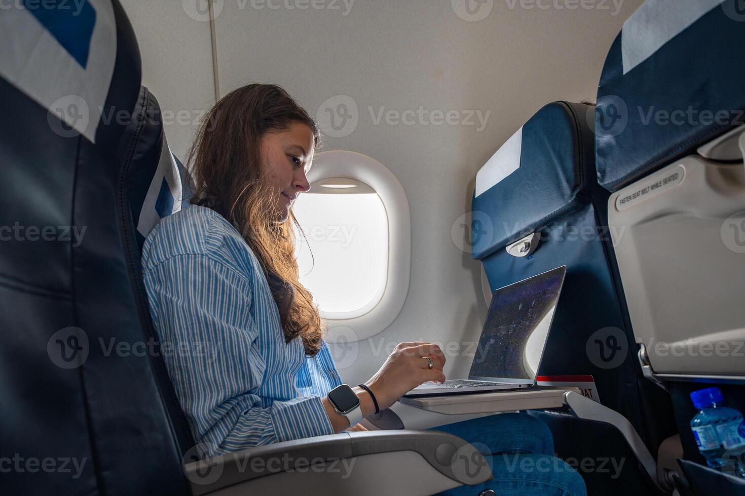 Young woman working on a laptop during a flight, seated by the airplane window. Remote work, travel productivity, and digital lifestyle on the go. photo