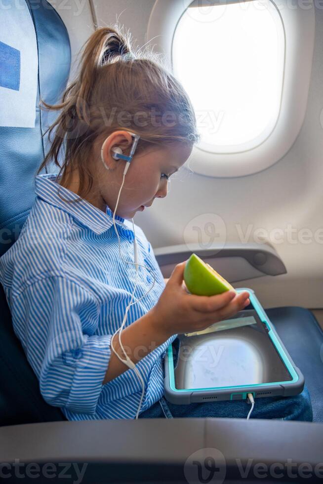 Child sitting in airplane seat during flight, wearing headphones, watching cartoons on a tablet and eating a green apple. Concept of children traveling, in-flight entertainment and healthy snack. photo