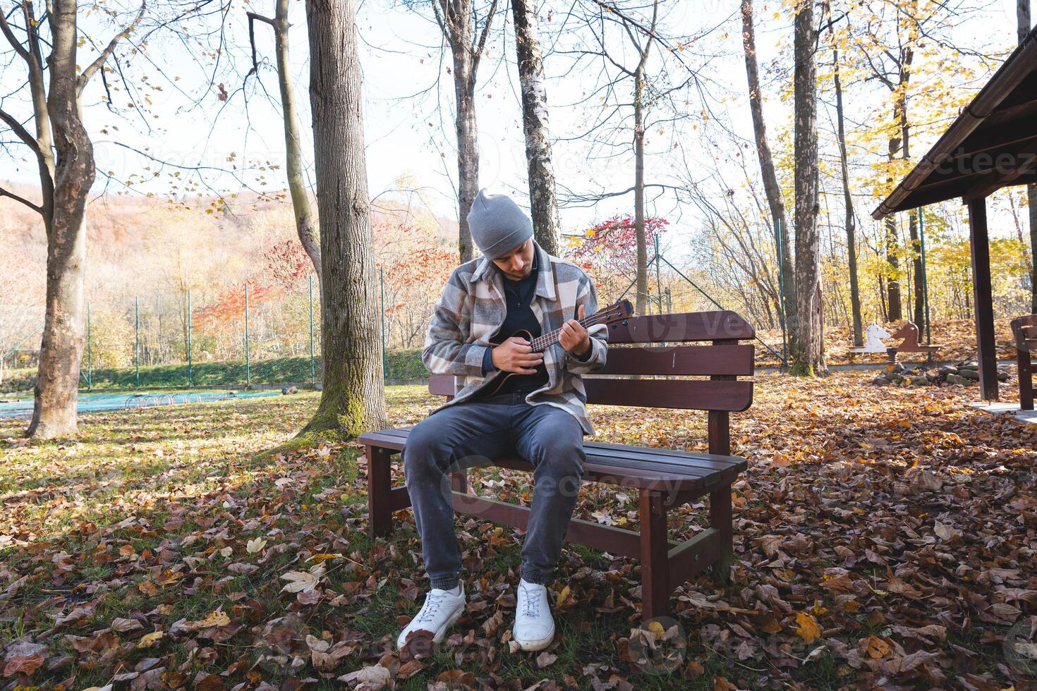 Enthusiast sits on a wooden bench in a park, playing the ukulele amidst autumn leaves. Surrounded by trees and bathed in soft sunlight, he enjoys a tranquil moment filled with creativity photo