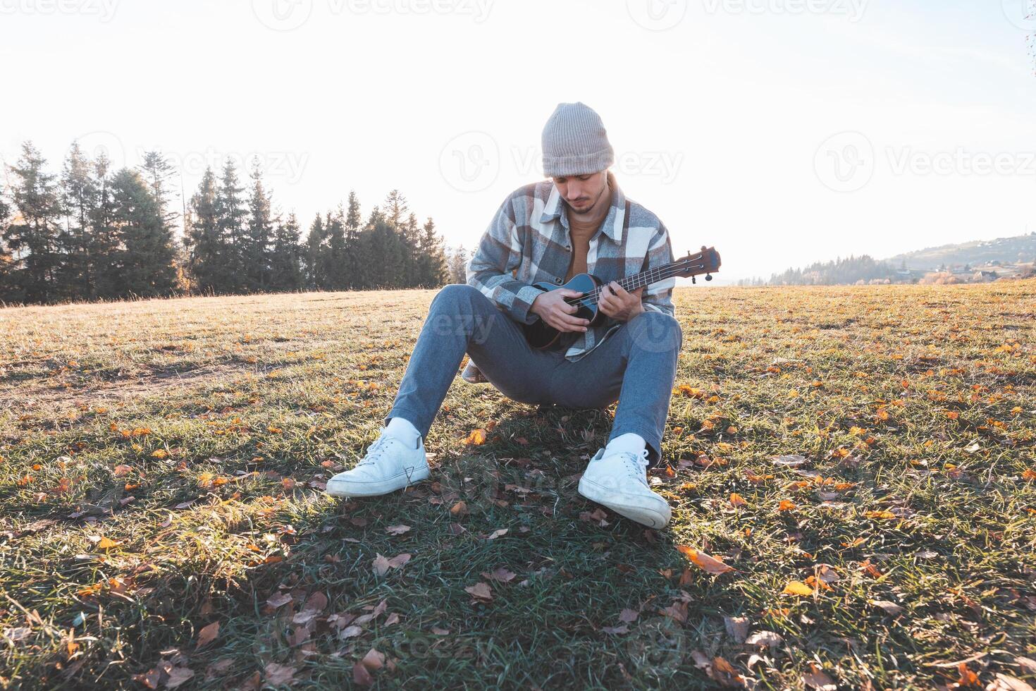 Musician sits on a grassy field in the warm autumn sunlight, smiling as he plays the ukulele. Surrounded by golden tones and distant trees, the moment reflects joy, creativity, and a deep connection photo