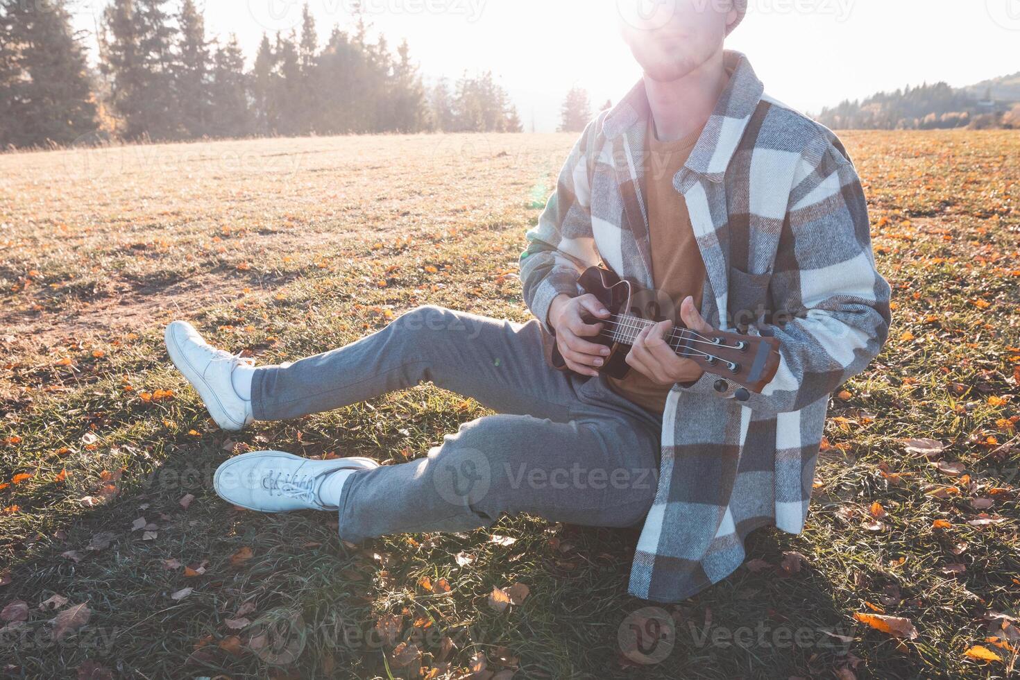 Beanie-wearing man sitting cross legged plays ukulele in the field, surrounded by trees and bathed in golden afternoon light photo