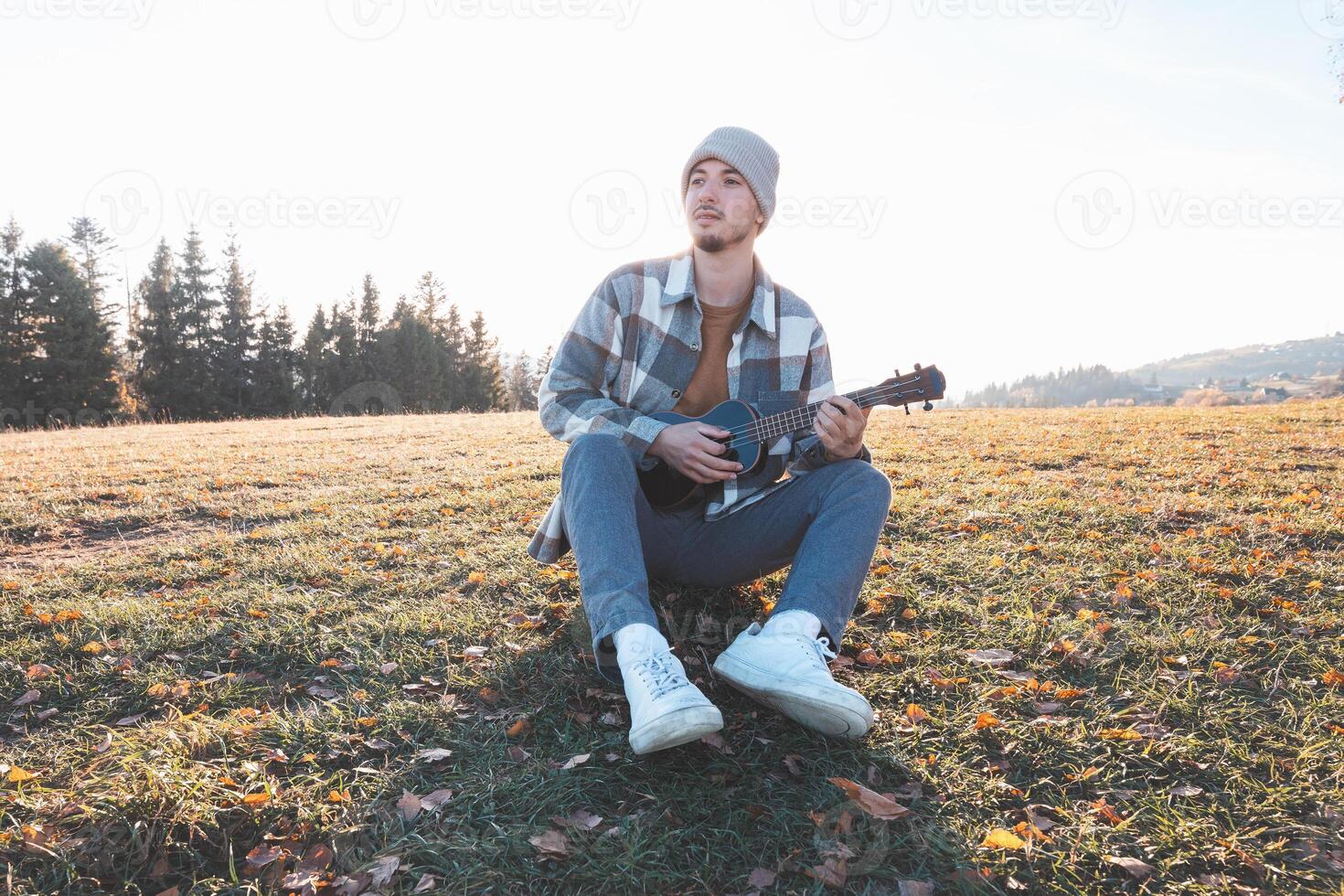 Happy traveler sits on a grassy field in the warm autumn sunlight, smiling as he plays the ukulele. Surrounded by golden tones and distant trees, the moment reflects joy, creativity photo