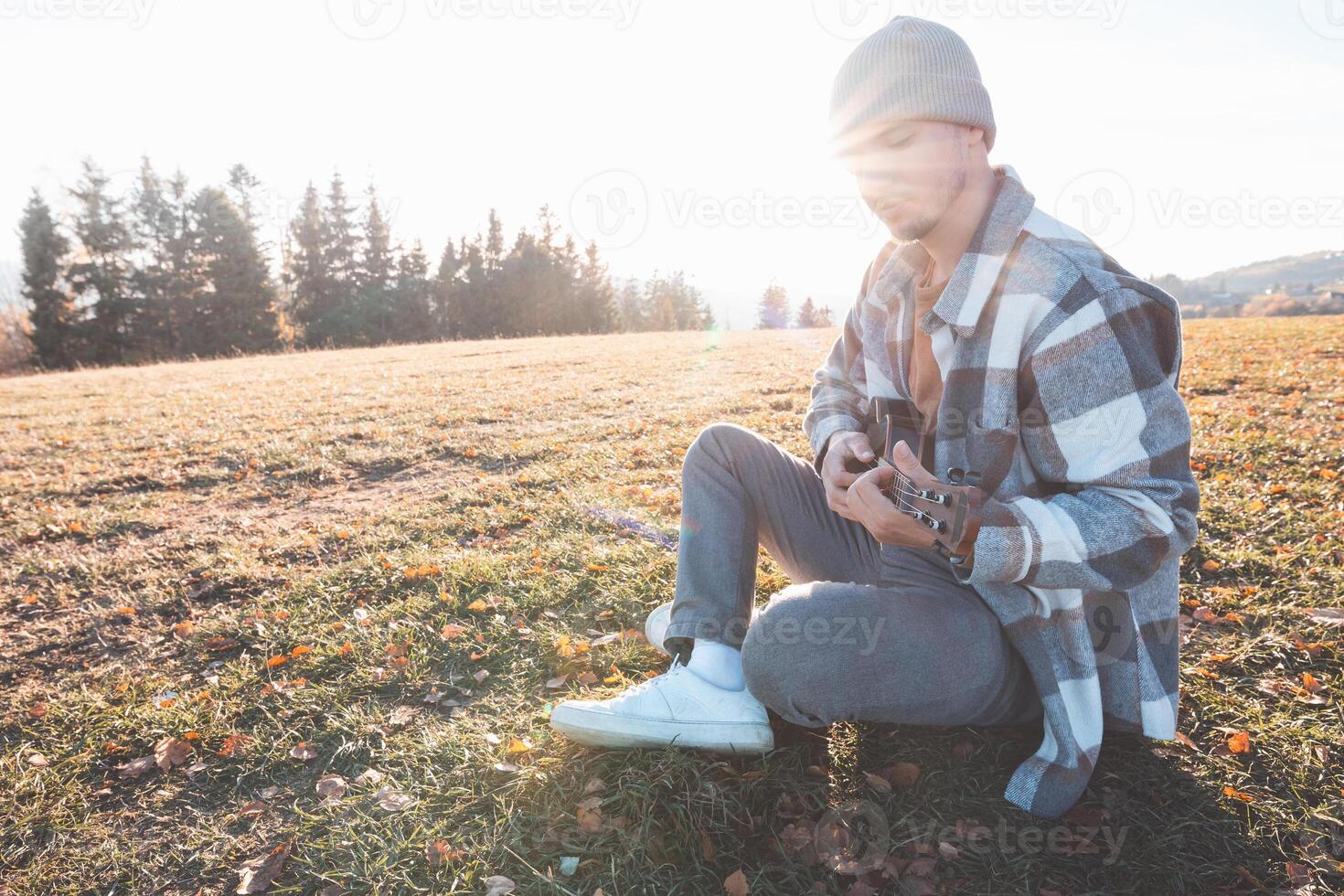 A solo ukulele player finds inspiration in nature while sitting quietly in the field with the sun lighting the landscape and trees all around photo