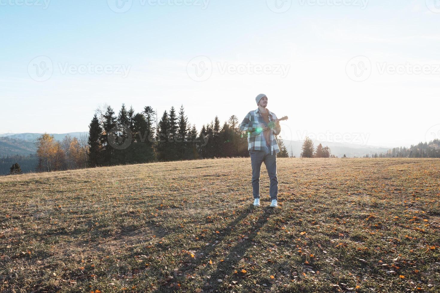 Traveler stands in an open field during a sunny autumn day, holding a ukulele. Surrounded by hills and trees, the serene scene reflects freedom, creativity, and a connection to nature photo
