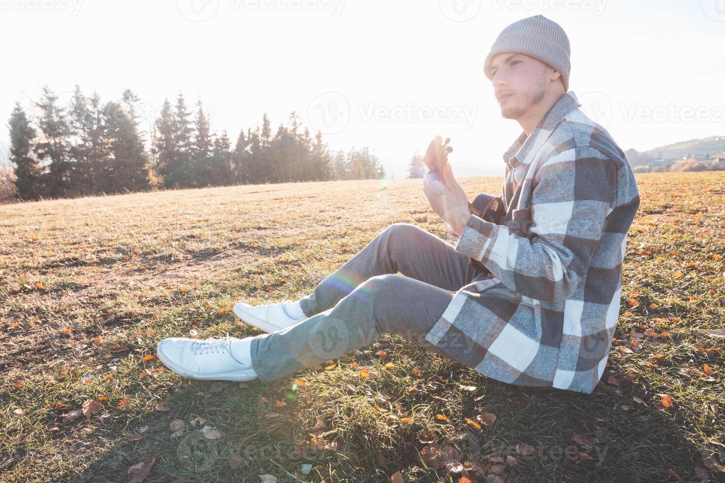 Young man in a beanie enjoys playing ukulele while sitting in a quiet open field, surrounded by nature and warmed by soft afternoon sunlight photo
