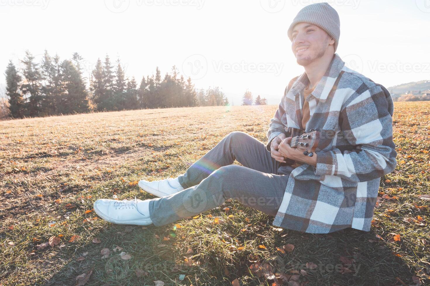 Casual man strumming ukulele outdoors in a wide open field on a sunny day with forest in the background and autumn leaves underfoot photo