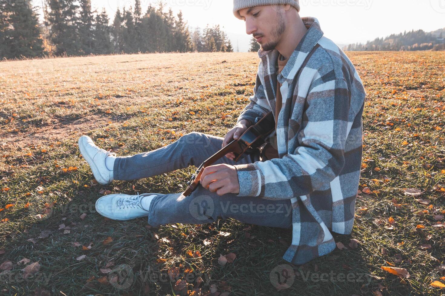 Casual man relaxes on autumn leaves while playing ukulele alone in a scenic field with forest edge and late sunlight in the background photo