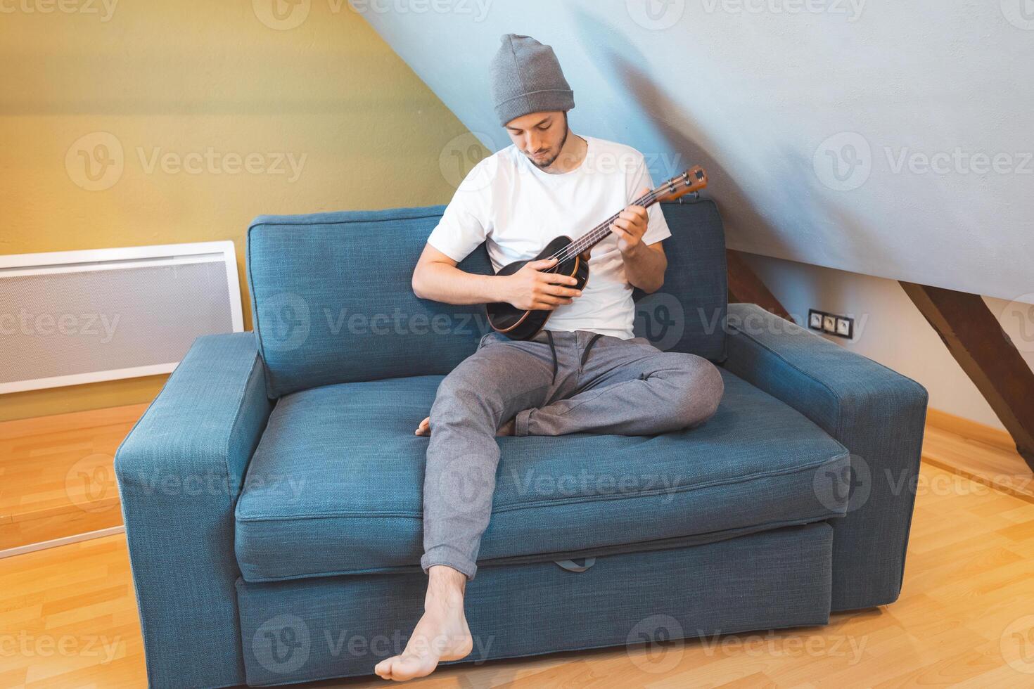 Man with beanie plays ukulele on a sofa in natural light photo