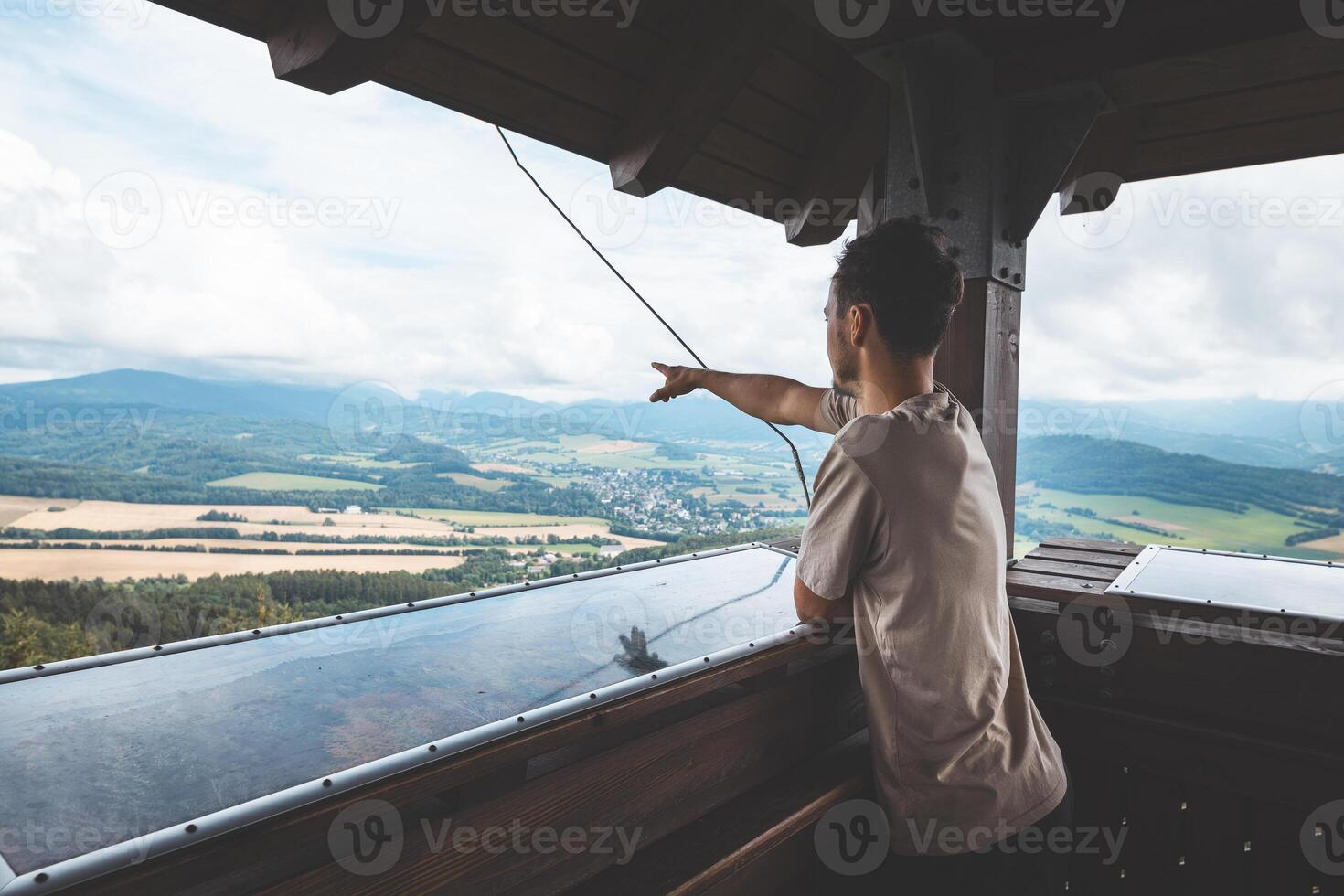 Finger points at a weathered orientation board on a viewing platform, labeling peaks of the Jeseniky Mountains unfolding across lush forested hills photo