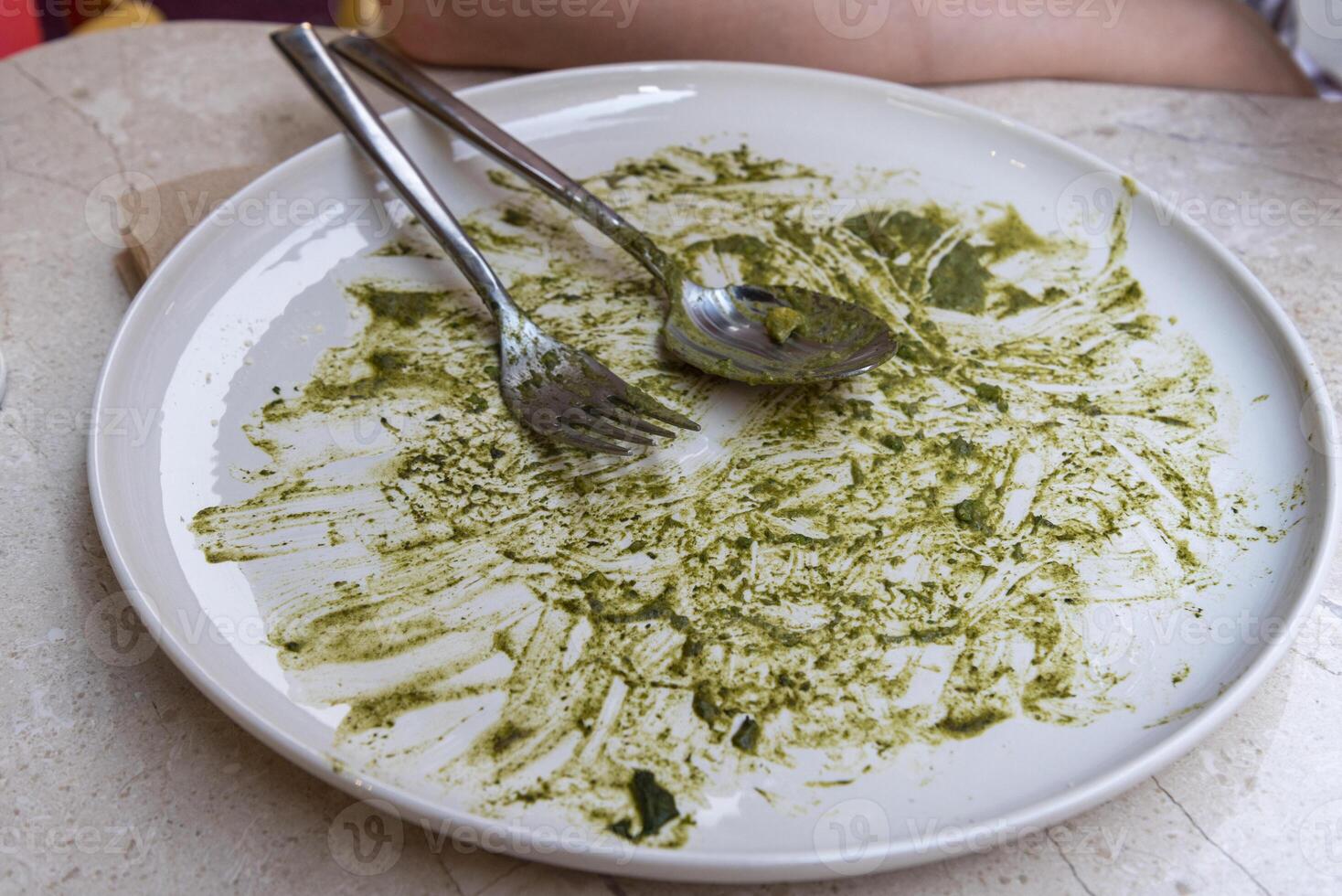 Empty plate with traces of green sauce and utensils resting on it at a dining table photo