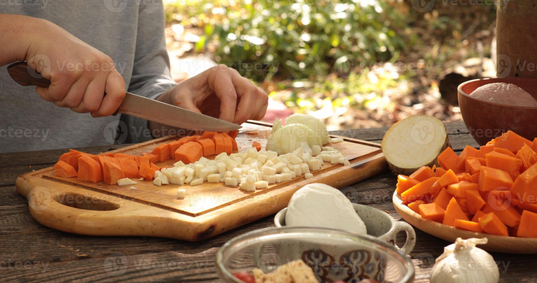 Chopping vegetables outdoors during sunny afternoon, focusing on preparation for healthy meal, featuring carrots, onions, squash, and fresh herbs on wooden cutting board photo
