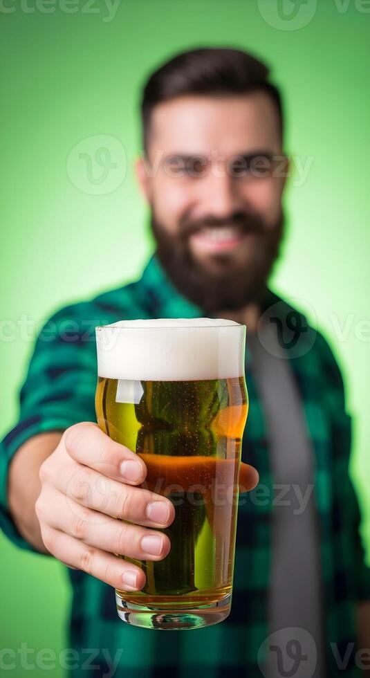 A man with a beard holds a glass of fresh light beer and stretches it to the camera on a green background. Vertical template for St. Patrick's Day, beer party, Oktoberfest and beer factory advertising photo
