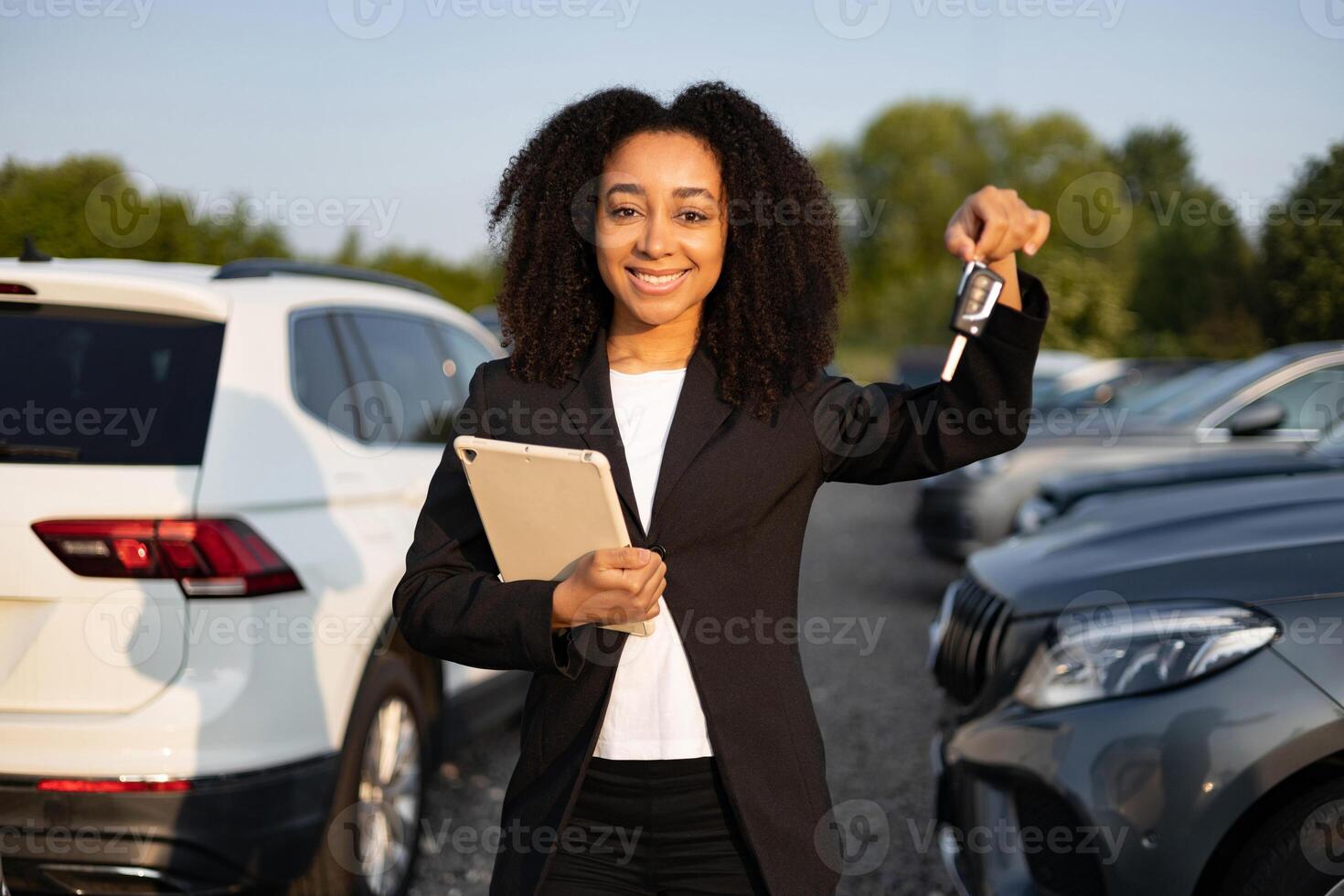 An African American woman, car salesperson, smiles while holding car keys and a tablet with vehicles visible in the background. photo