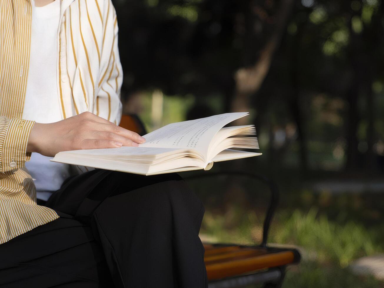 Woman Reading Open Book in Park On Bench Under Tree Wearing White Shirt and Jacket photo