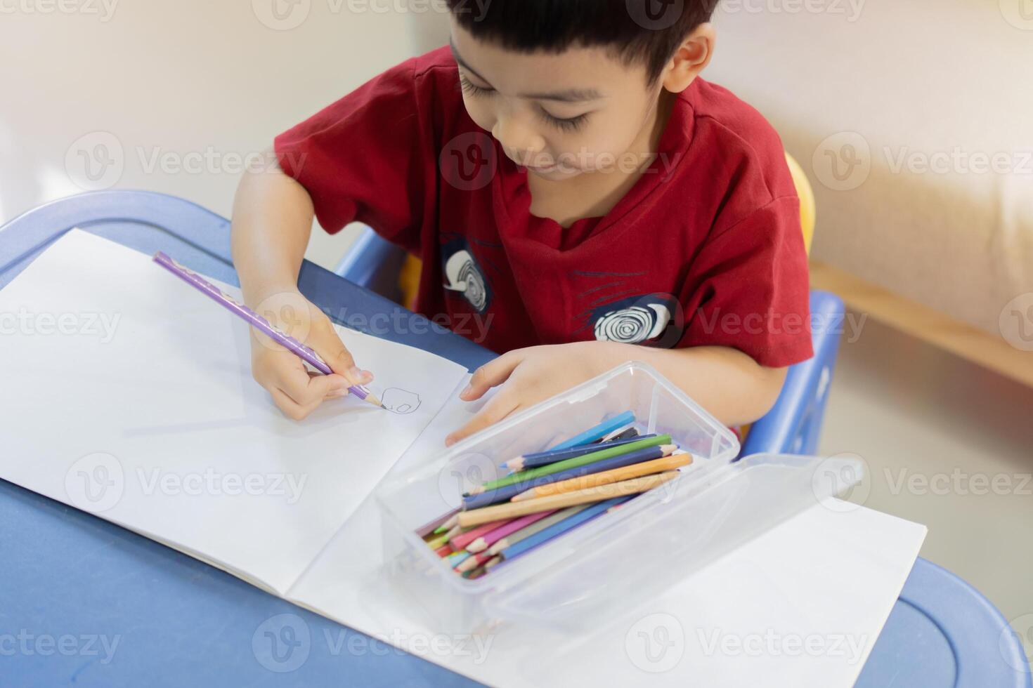 Cute little Asian boy drawing with colorful pencils, sitting on desk at home, alone. photo