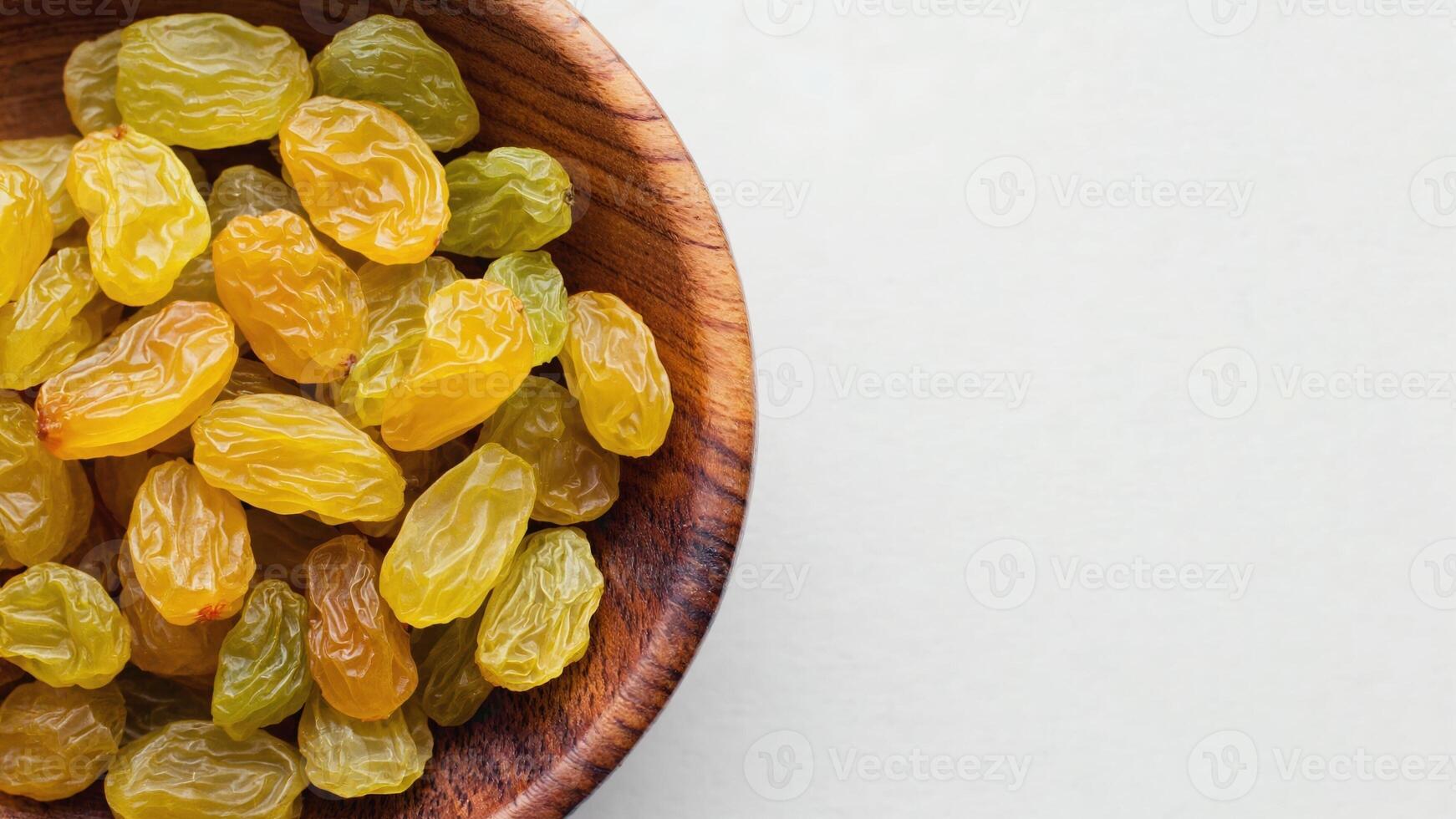 a wooden bowl full of golden raisins, located on the left on a white background. The raisins are slightly poured over the edge, creating an appetizing and natural look. photo