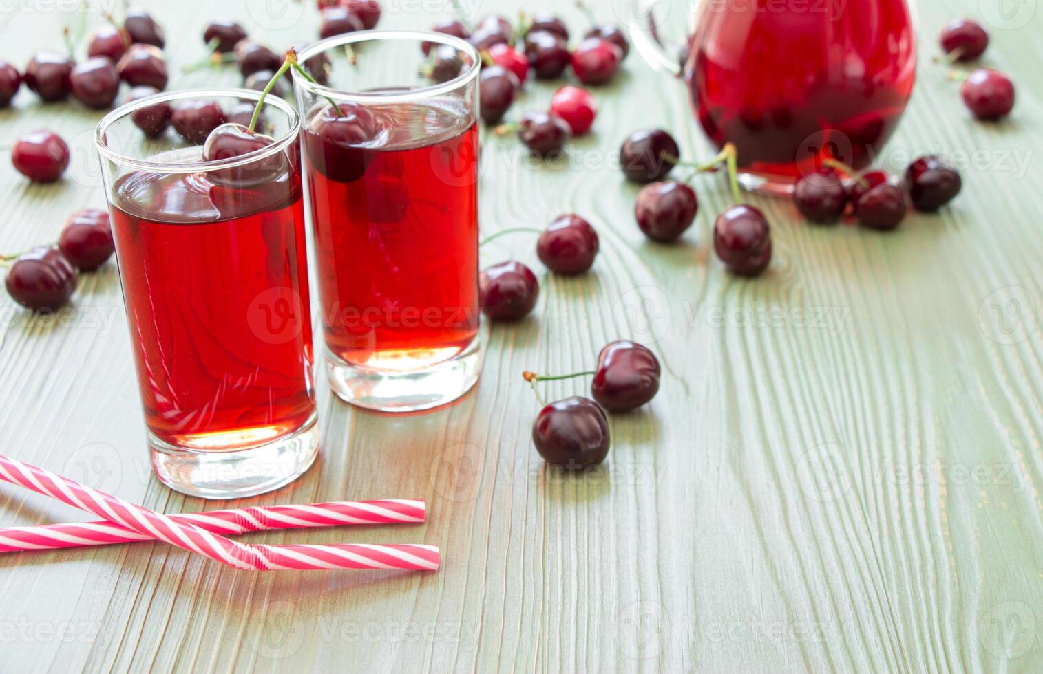 Left 2 glasses of juice, straws, scattered cherries, jar of cherry juice, on right empty space for text on green background. Cherries, cherry juice and empty space. Horizontal. photo