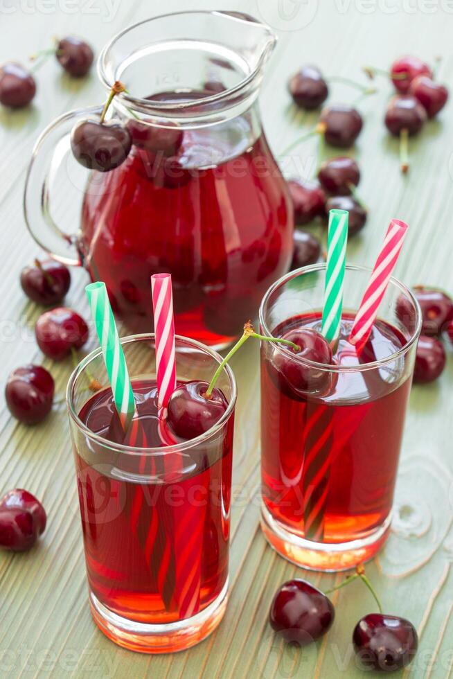 Glasses of cherry juice with straws in the foreground and in the background a jar full of cherry juice. 2 cherry juices. Vertical. Close-up. Daylight. photo