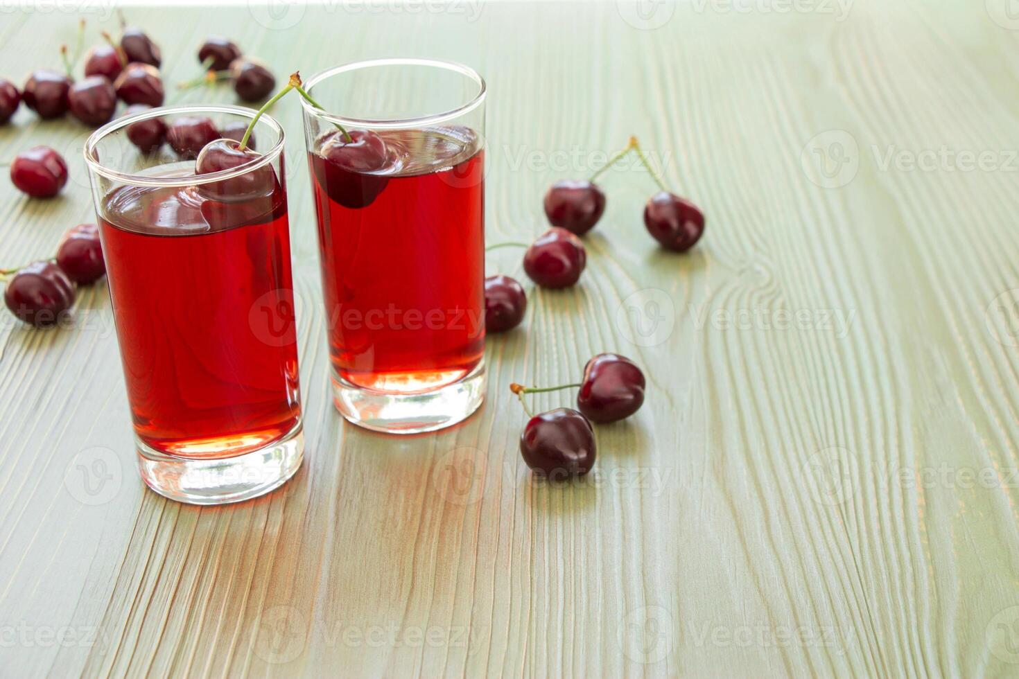 Two clear glasses filled with vibrant cherry juice sit on a light wooden table. Fresh cherries are scattered around, adding a colorful touch to the scene in a bright environment. photo