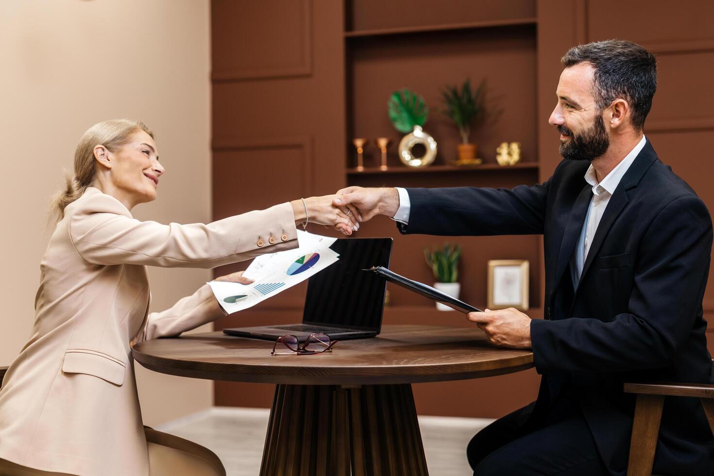 Business professionals engaged in a handshake at a round table, exchanging documents and charts, symbolizing collaboration and strategic planning in a modern office environment photo