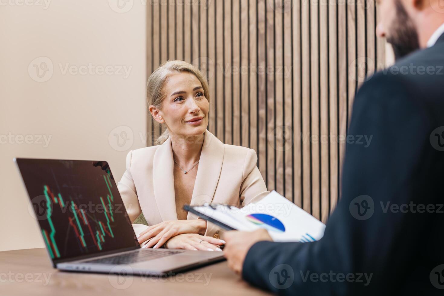 Businesswoman in a beige suit engaged in a meeting, analyzing financial data on a laptop, while a colleague presents a report with charts and graphs in a modern office setting photo