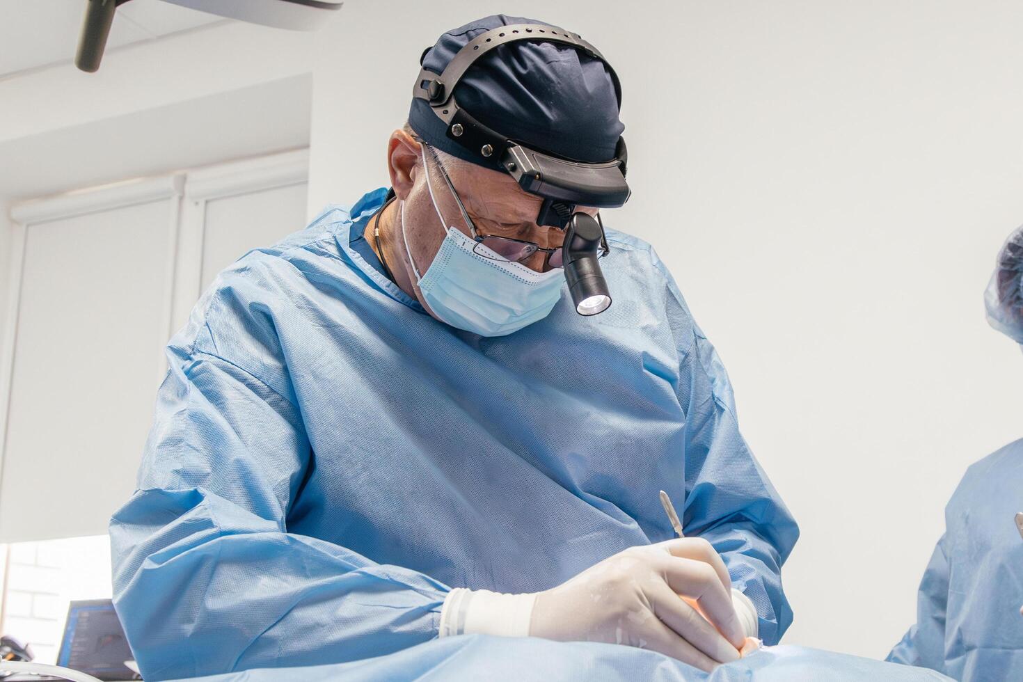 Male surgeon in blue scrubs and surgical mask is performing an operation with precision, using specialized tools in a bright, sterile medical environment, showcasing dedication to patient care photo