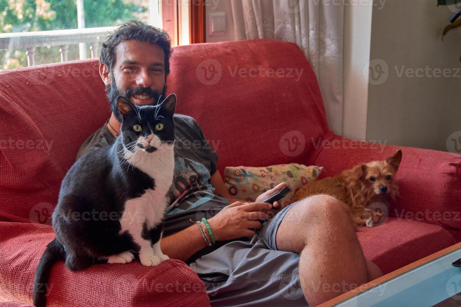 Bearded young man petting a small ginger dog while his bicolor cat rests nearby in a brightly lit living room photo