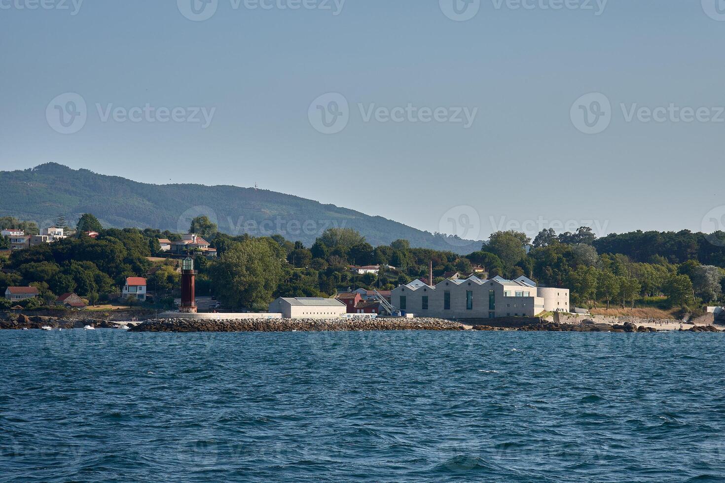 Panoramic view of the Museo do Mar and lighthouse in Vigo surrounded by trees with the sea in the foreground and a clear sky photo