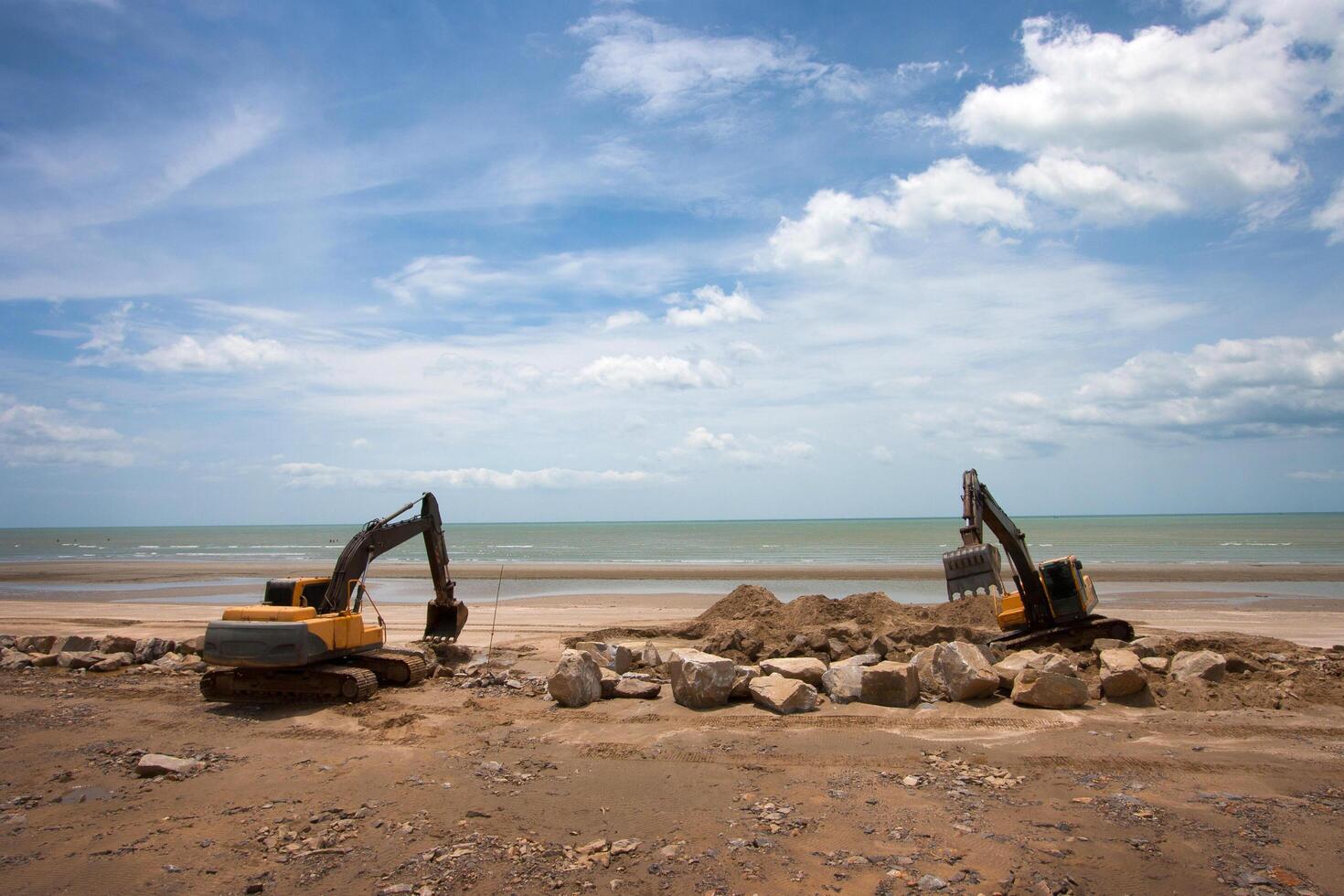 Two excavators on a sandy beach, surrounded by rocks, under a bright, cloudy sky, with the ocean in the background. photo