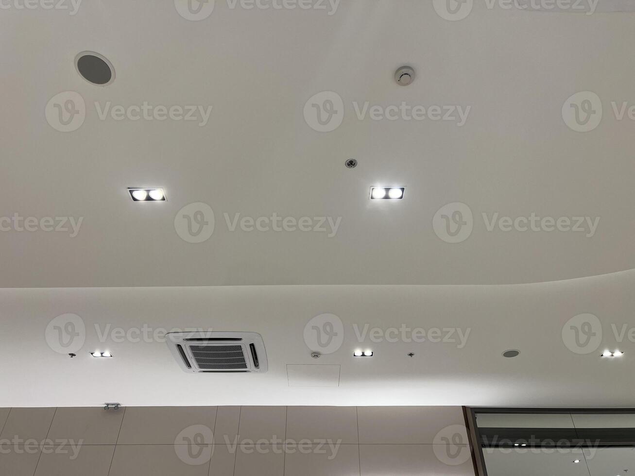 Functional and modern white ceiling with recessed lighting, air conditioner, security camera, smoke detector, and speaker creating secure and comfortable atmosphere photo