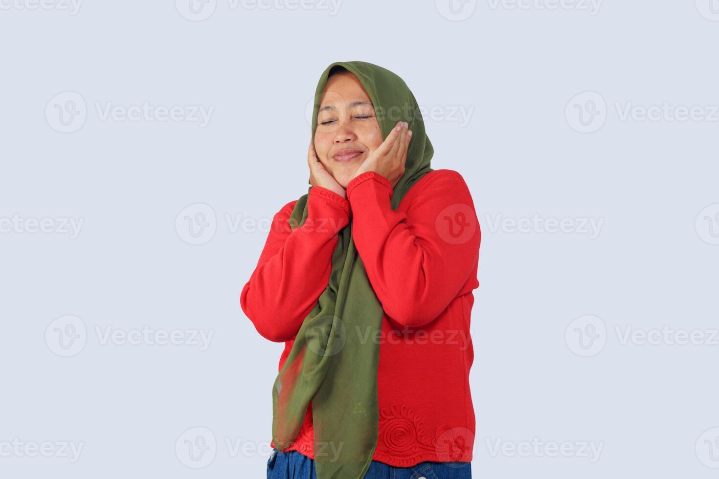 Joyful muslim woman in hijab smiling with eyes closed and hands on cheeks, expressing happiness and satisfaction, isolated on plain background photo
