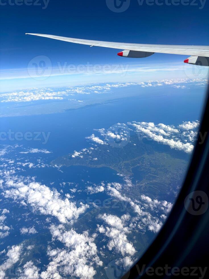 An aerial view from an airplane window. The airplane wing frames a landscape of forested mountains, deep blue sea, and scattered clouds beneath a bright sky. Scenic or business flight travel photo. photo